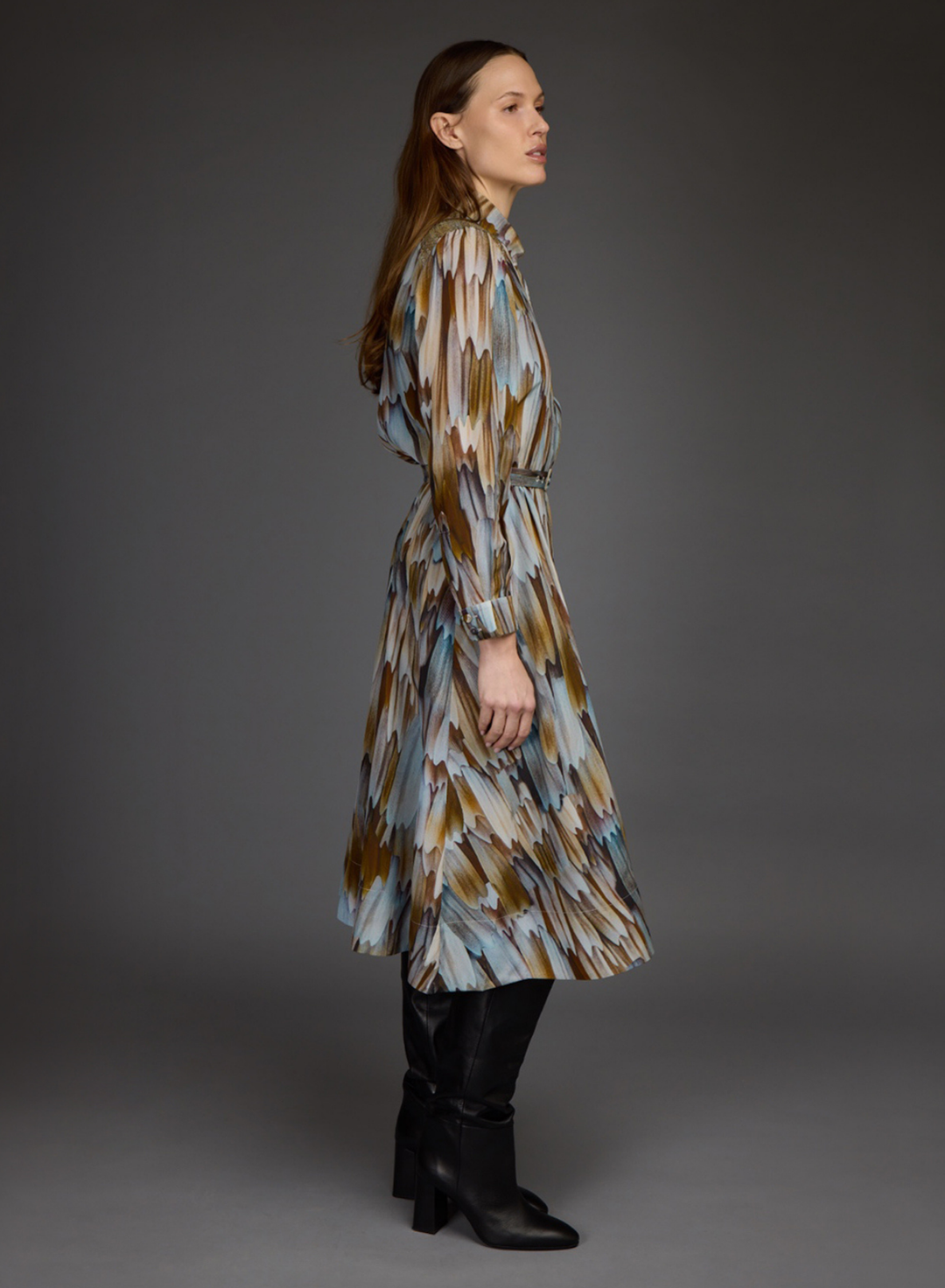 A woman stands in profile wearing a long-sleeve, knee-length dress with an abstract brushstroke pattern in beige, brown, light blue, and white, paired with black knee-high boots against a dark gray studio background.
