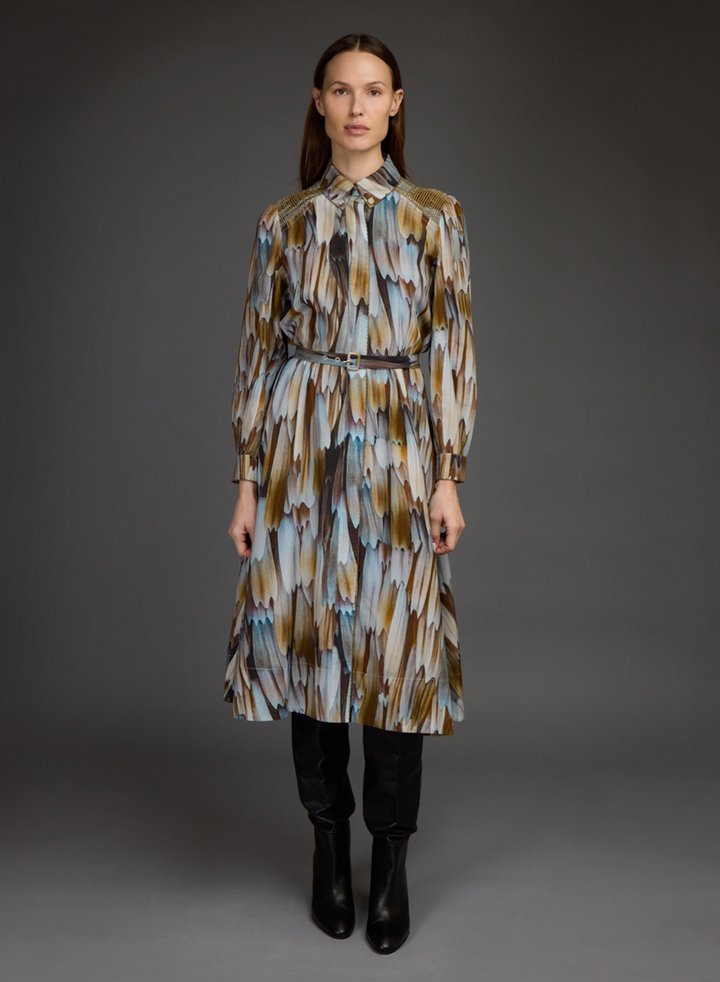 A model stands against a dark gray studio background wearing a long-sleeved midi shirt dress with a brown, beige, and light blue feather-like abstract print, styled with a thin dark belt and black ankle boots.