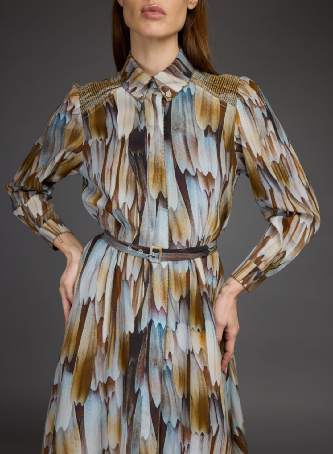 A woman wearing a long-sleeve collared dress with an abstract feather-like print in brown, beige, white, and light blue, cinched at the waist with a thin belt.