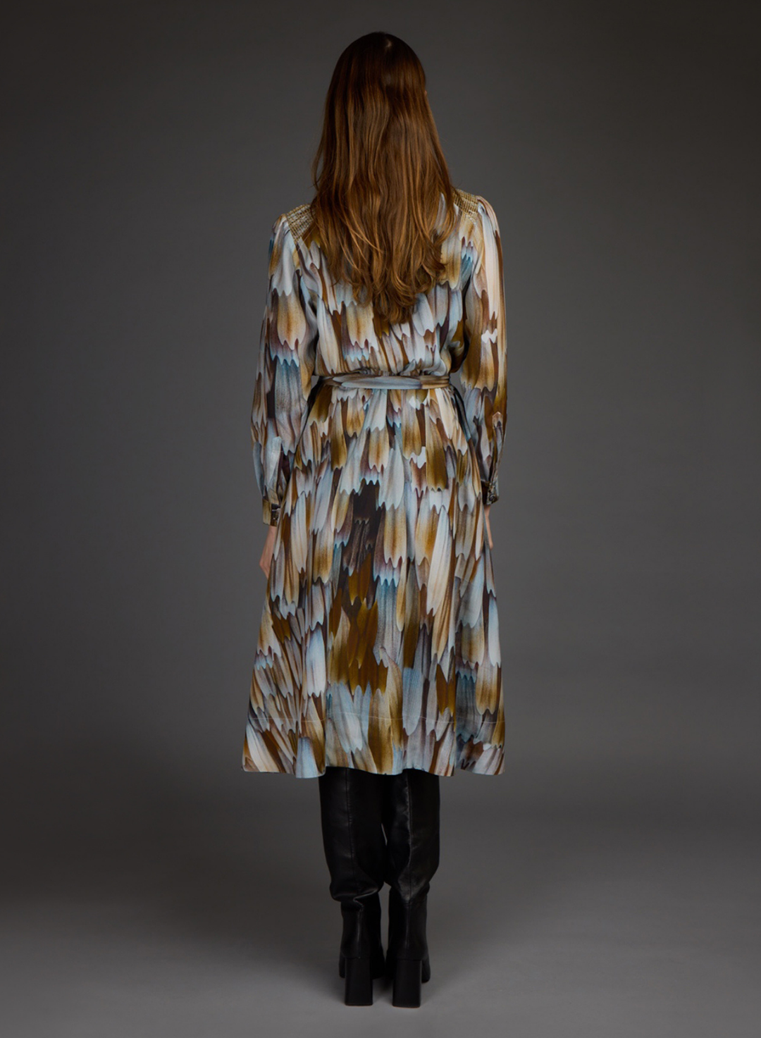 A woman viewed from the back wearing a long belted wrap coat with an abstract ikat-style pattern in brown, beige, and light blue, paired with black boots against a dark gray studio background.