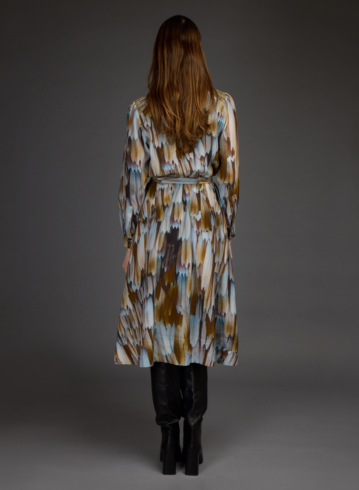 A woman viewed from the back wearing a long belted wrap coat with an abstract ikat-style pattern in brown, beige, and light blue, paired with black boots against a dark gray studio background.