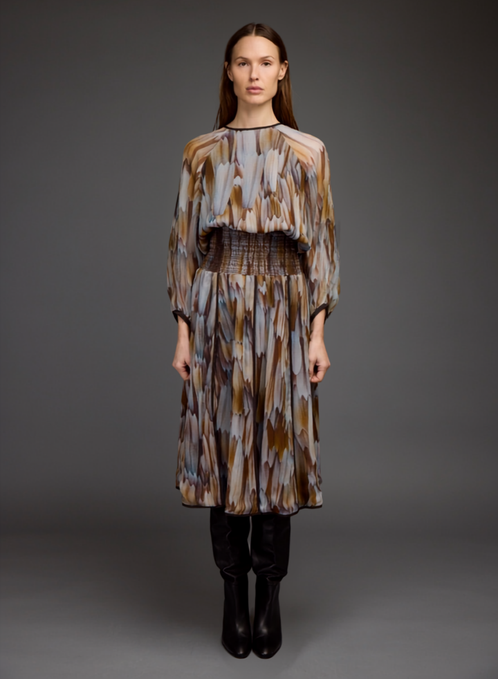 A model stands against a dark gray studio background wearing a long-sleeved midi dress with an abstract brushstroke pattern in brown, beige, and gray, paired with black ankle boots.