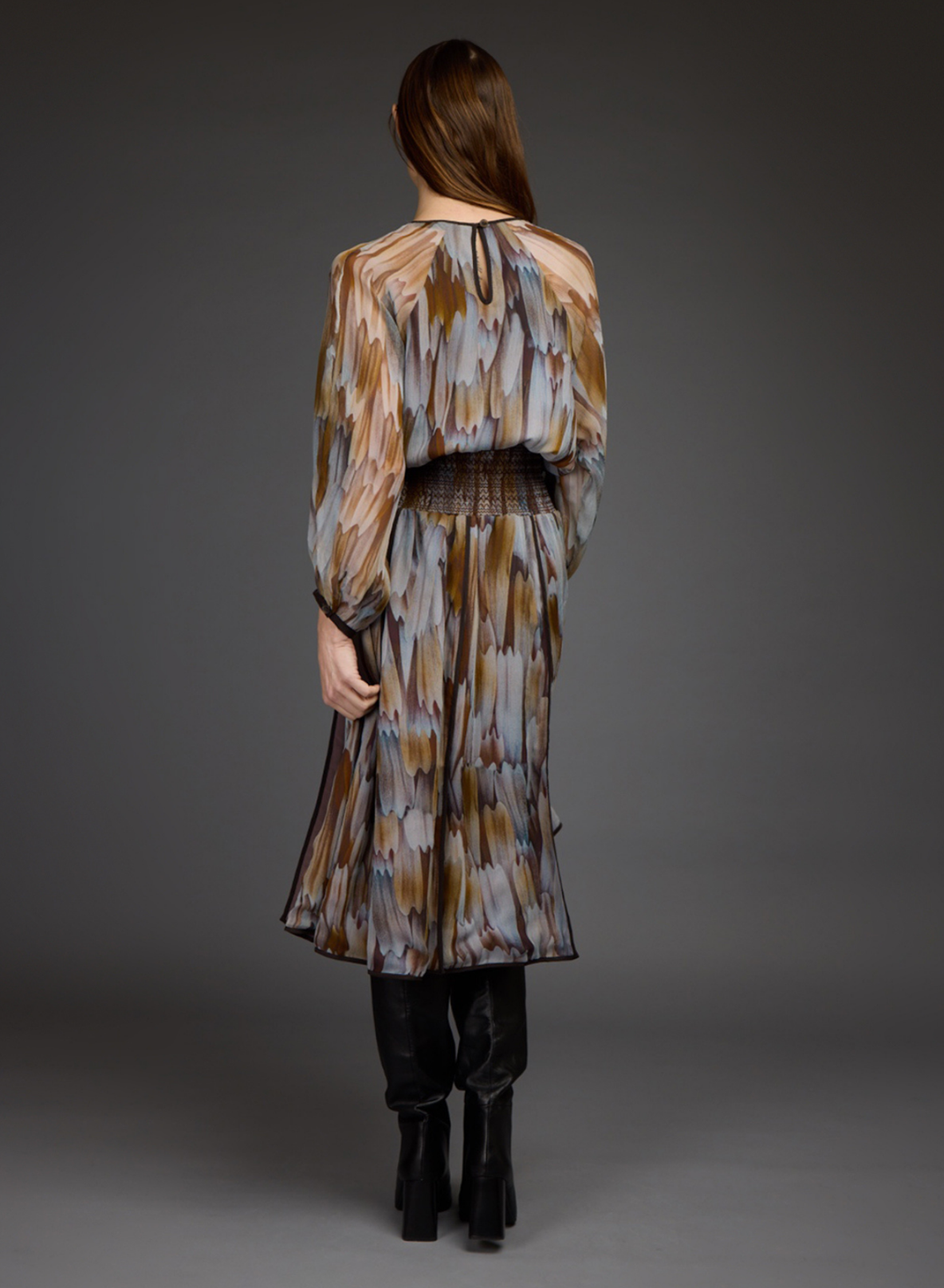 A woman viewed from behind wearing a long-sleeved pleated midi dress with an abstract brown, beige, and gray print, paired with black knee-high boots, against a dark gray studio background.