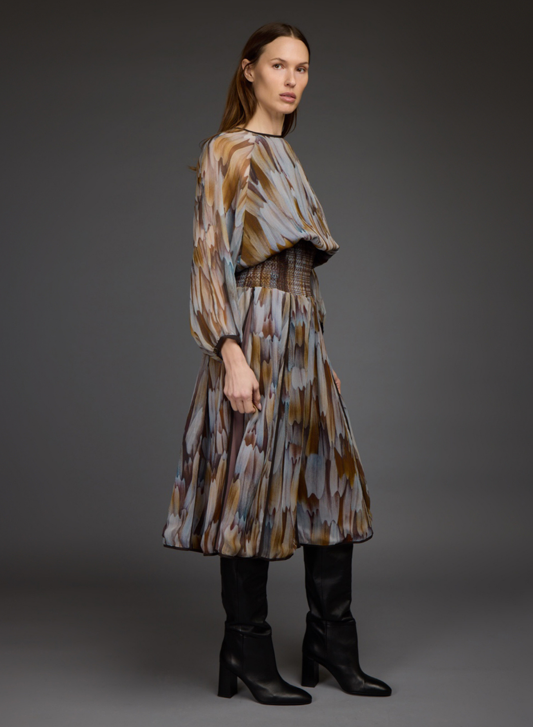 A model wearing a long-sleeved midi dress with a brown, beige, and gray abstract pattern, styled with black leather knee-high boots against a dark gray studio background.