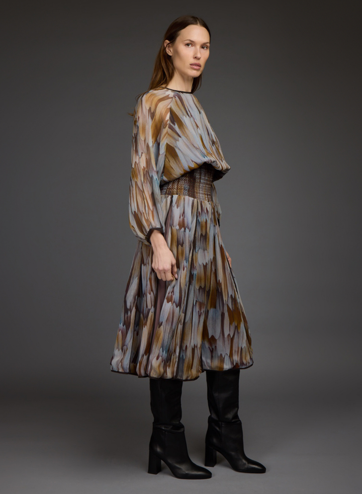A model wearing a long-sleeved midi dress with a brown, beige, and gray abstract pattern, styled with black leather knee-high boots against a dark gray studio background.