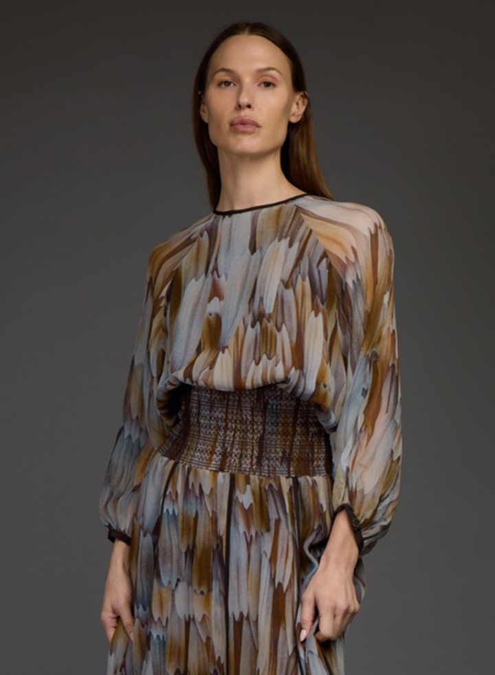 A woman modeling a long-sleeved midi dress with an abstract, painterly brushstroke pattern in brown, beige, gray, and muted blue tones, featuring a smocked waistband.
