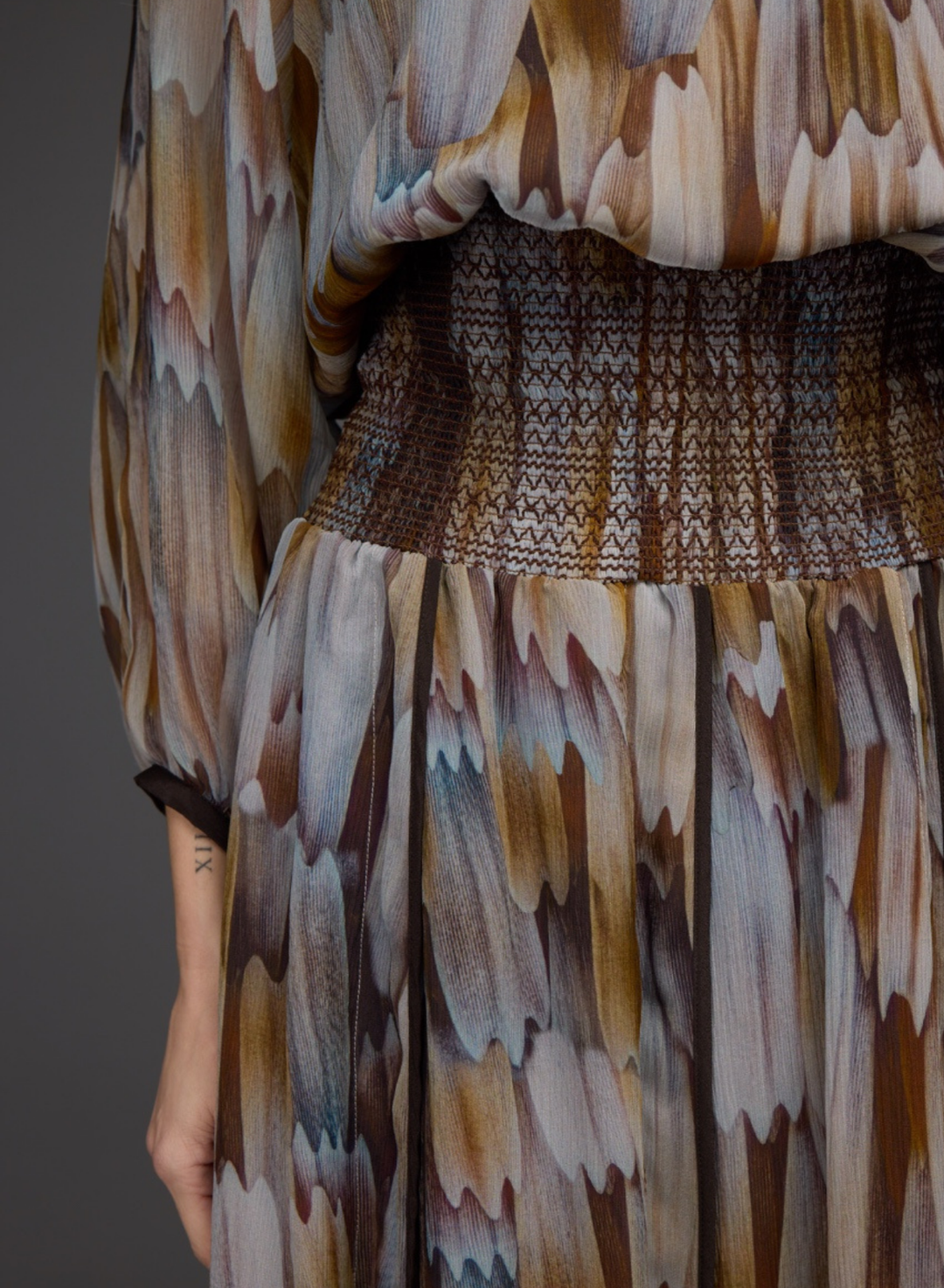 A close-up of a model wearing a long-sleeved dress with a brown, beige, and gray abstract patterned print, featuring a smocked waistband.