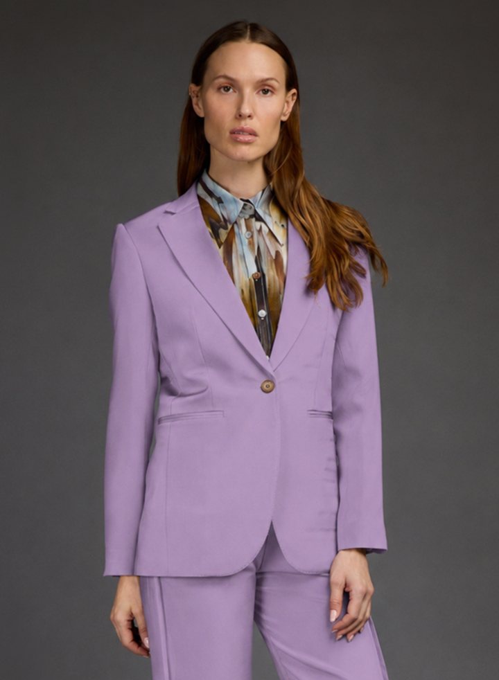 A woman modeling a lavender purple tailored blazer and matching trousers, worn over a patterned blue, brown, and beige shirt, against a dark gray studio background.