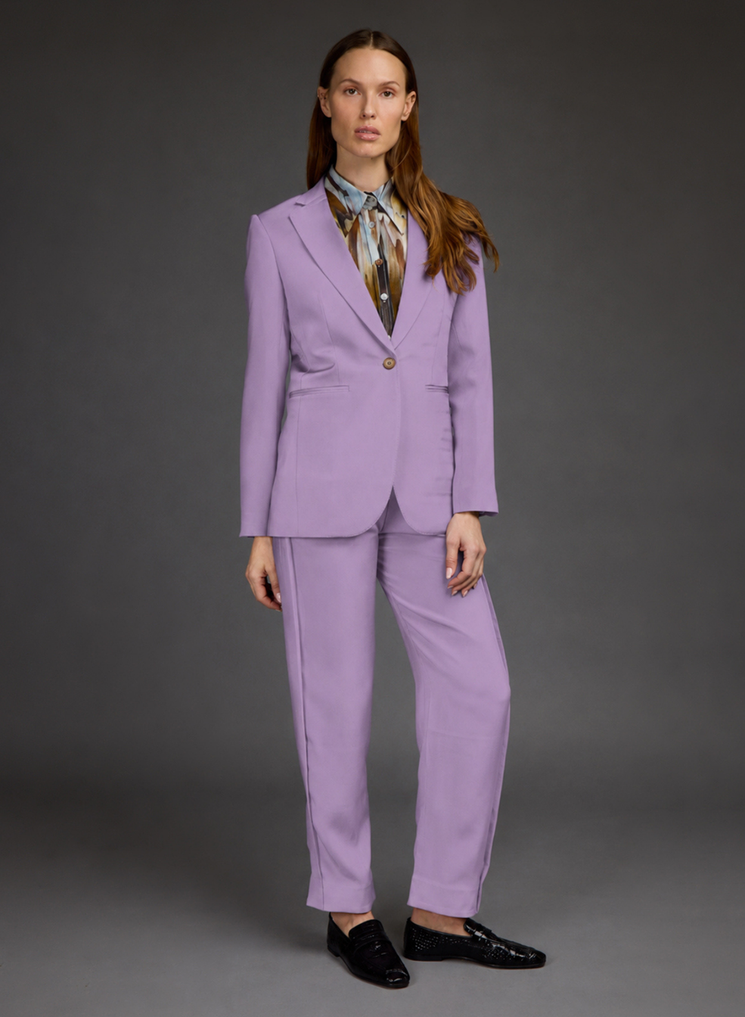 A model stands against a dark gray studio background wearing a lavender-purple tailored suit with a single-breasted blazer and matching trousers, styled with a patterned neck scarf and black loafers.
