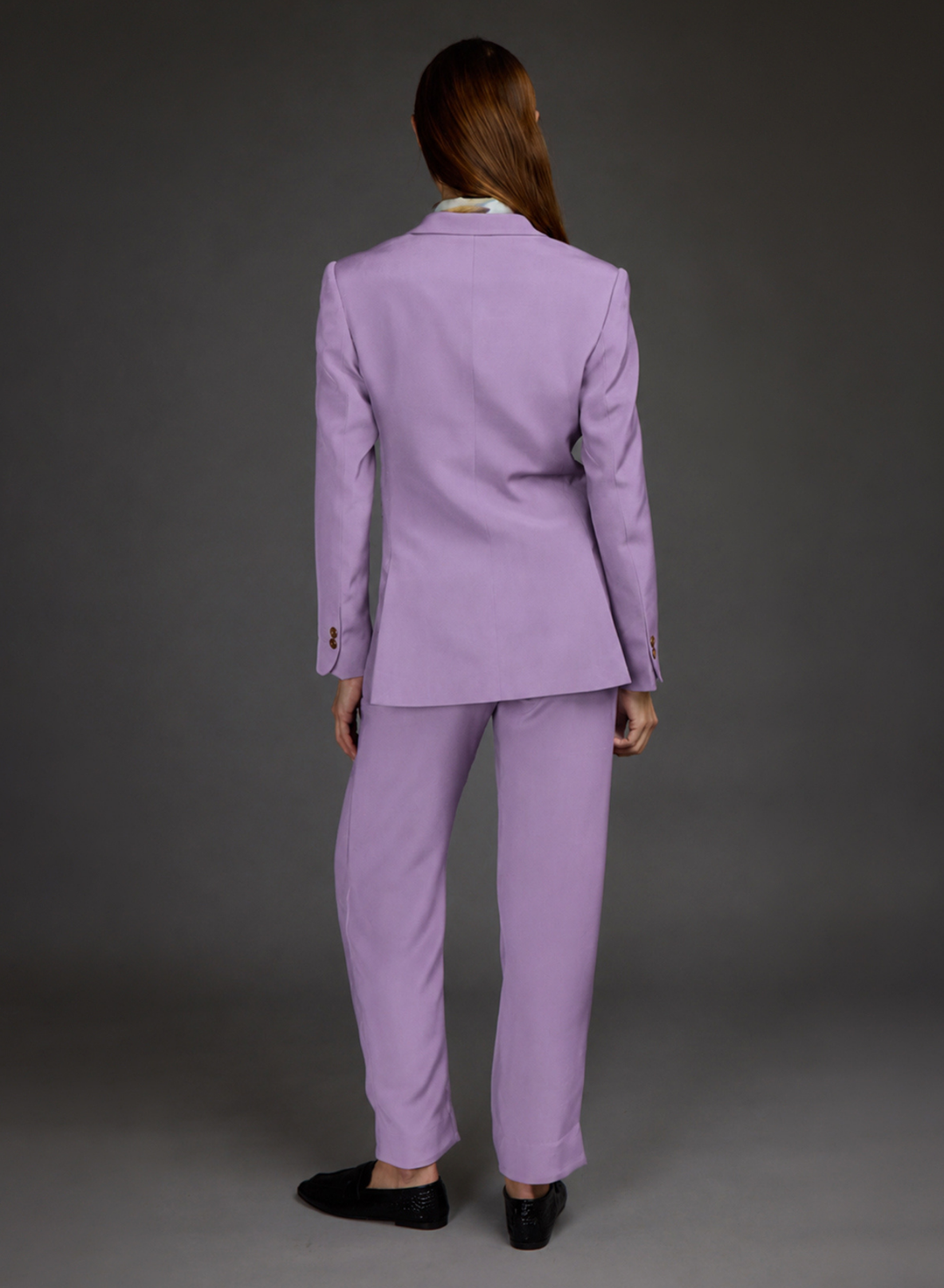 A model shown from the back wearing a lavender purple tailored suit, consisting of a blazer and matching trousers, paired with black loafers against a dark gray studio background.