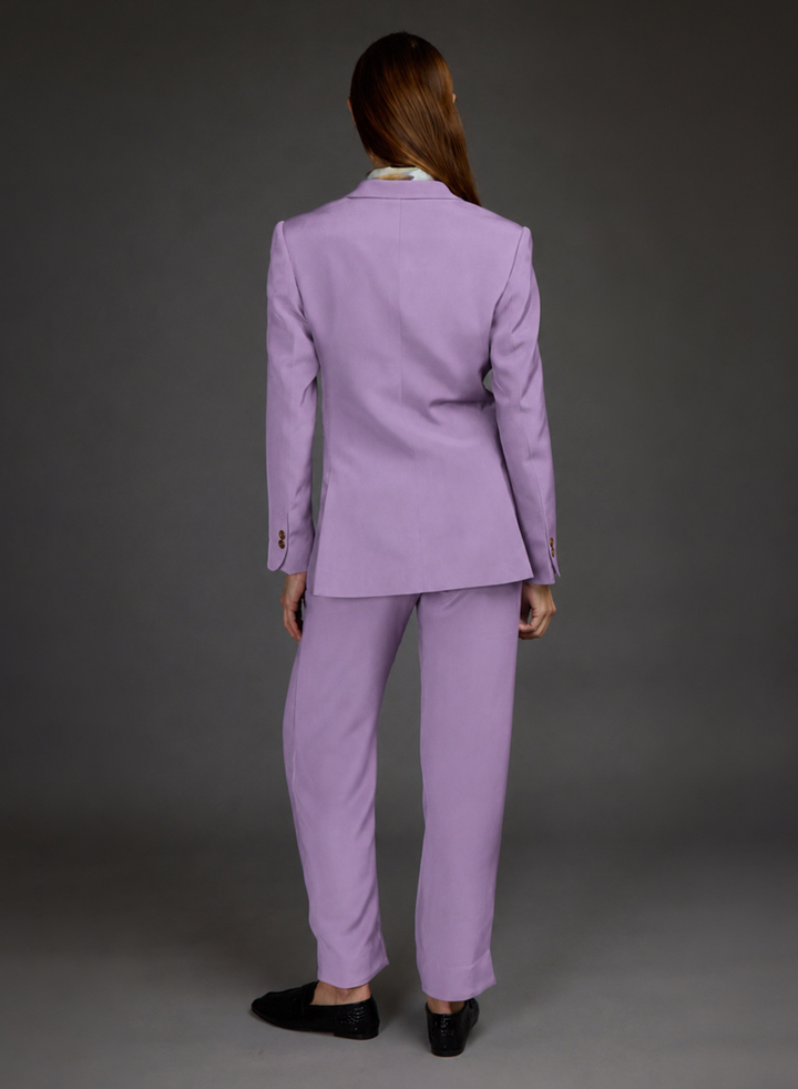 A model shown from the back wearing a lavender purple tailored suit, consisting of a blazer and matching trousers, paired with black loafers against a dark gray studio background.