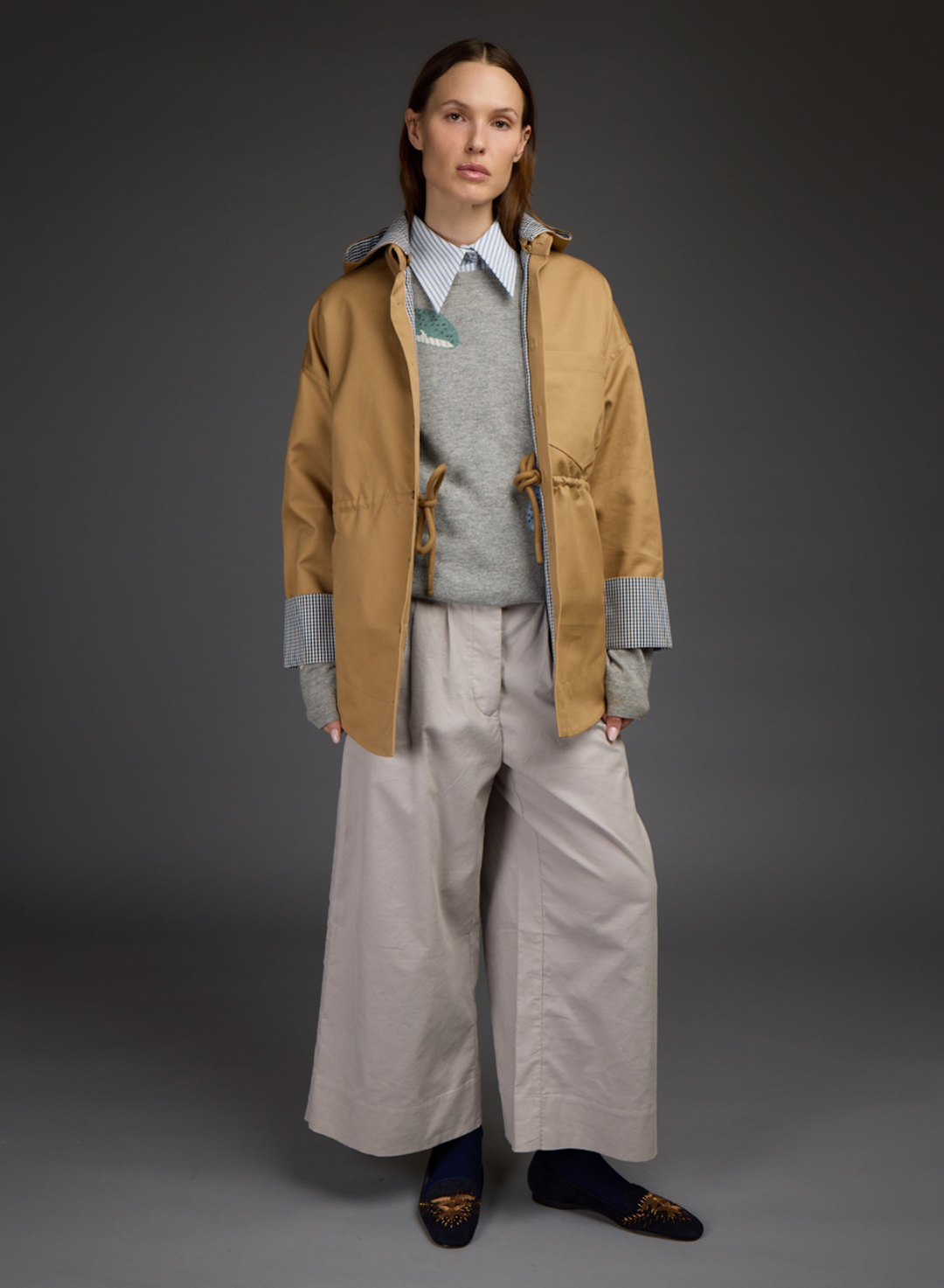 A model stands against a dark gray studio background wearing a tan hooded jacket over a gray sweater and light blue collared shirt, paired with wide-leg light beige trousers and dark loafers. All by Love Binetti. 