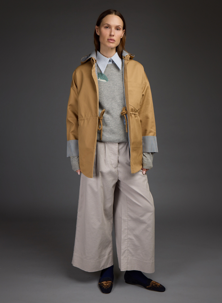 A model stands against a dark gray studio background wearing a tan hooded jacket over a gray sweater and light blue collared shirt, paired with wide-leg light beige trousers and dark loafers. All by Love Binetti. 