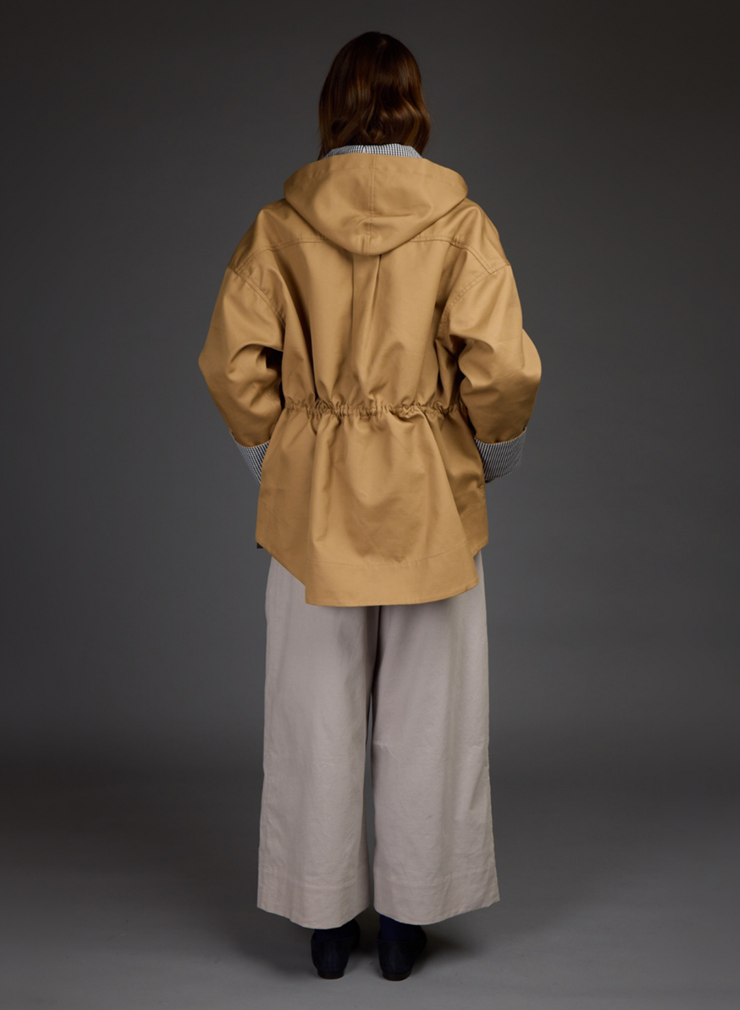 A model viewed from the back wearing a Love Binetti tan hooded jacket by with a drawstring waist, paired with wide-leg light gray trousers and black shoes, against a dark gray studio background.