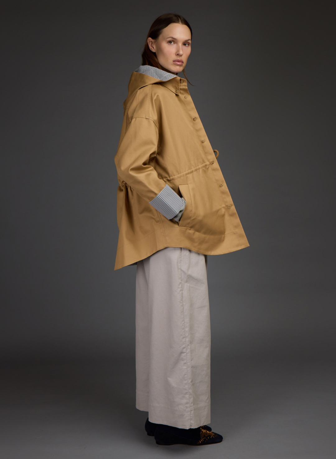 A model is shown from the side wearing a camel-colored hooded raincoat by Love Binetti over a light gray collared shirt and wide-leg light gray trousers, set against a dark gray studio background.
