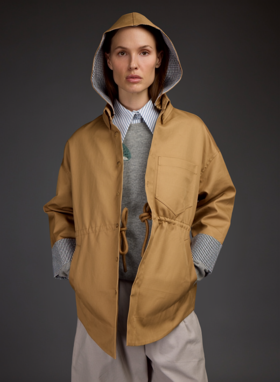 A model wearing a tan Love Binetti hooded rain jacket over a gray sweater with mushroom motif, striped shirt, and light trousers, photographed against a dark gray studio background.