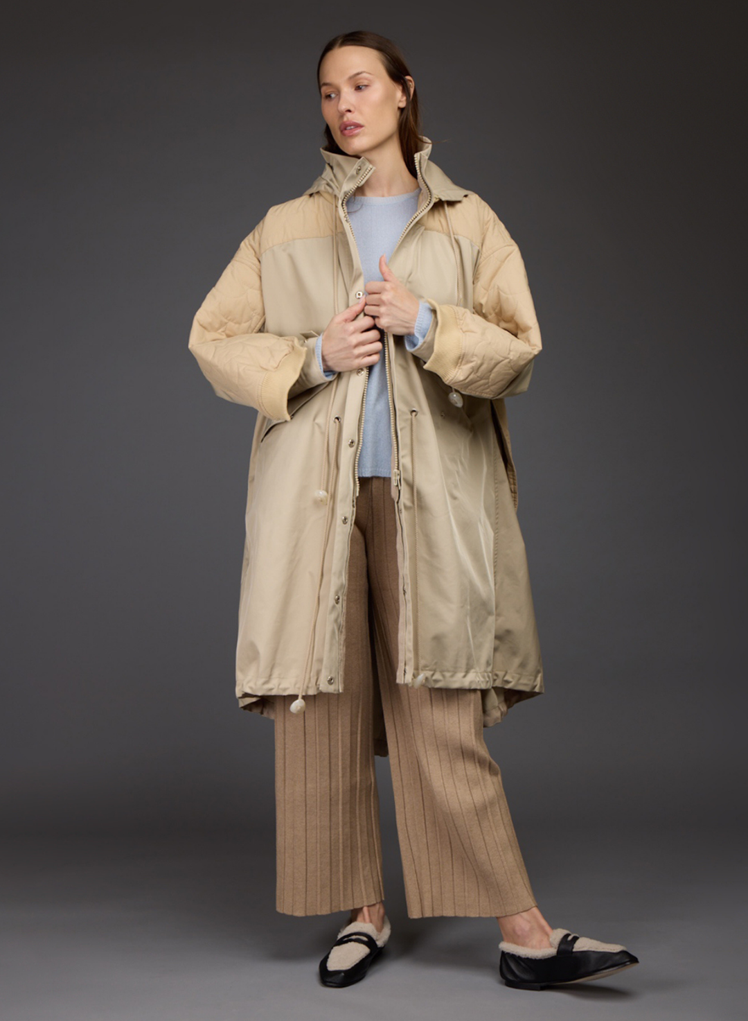 A model stands against a dark gray studio background wearing a long beige quilted puffer coat over a light blue top and beige pleated drawstring trousers, paired with black and beige loafers. All by Love Binetti. 