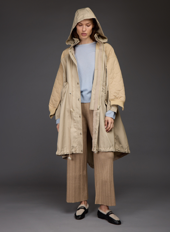 A model stands against a dark gray studio background wearing a long beige hooded quilted coat over a light blue sweater, beige pleated trousers, and black-and-white loafers. All by Love Binetti.