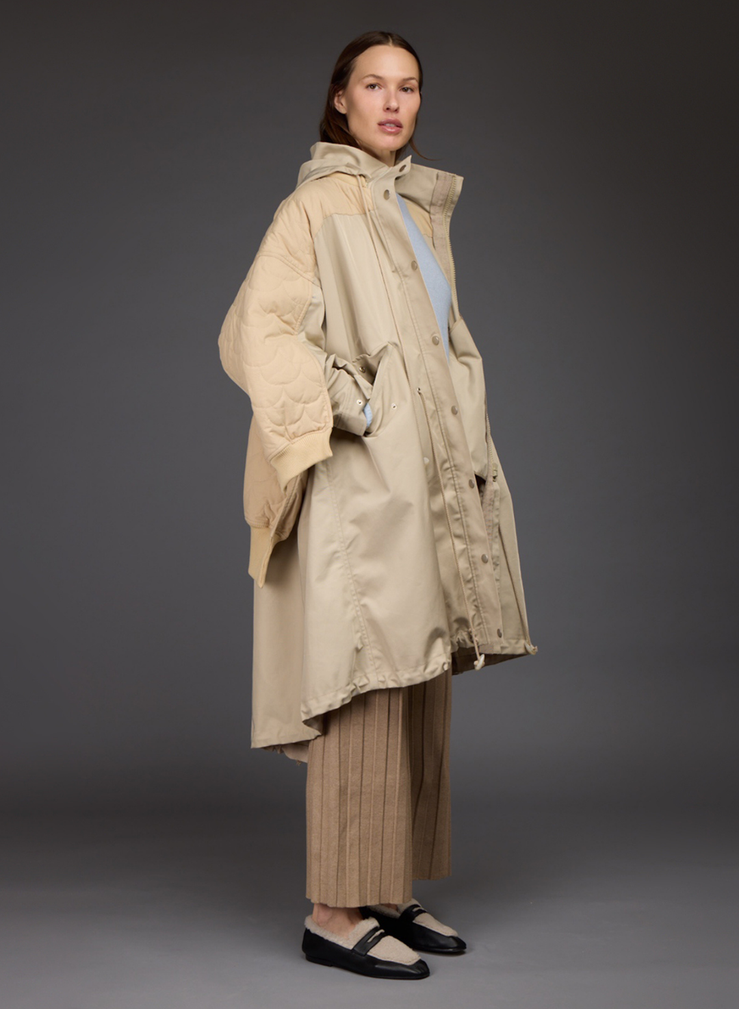 A model shown from the back wearing a beige quilted puffer jacket with a hood, a matching beige long coat, pleated beige trousers, and black and cream flats, set against a dark gray studio background.