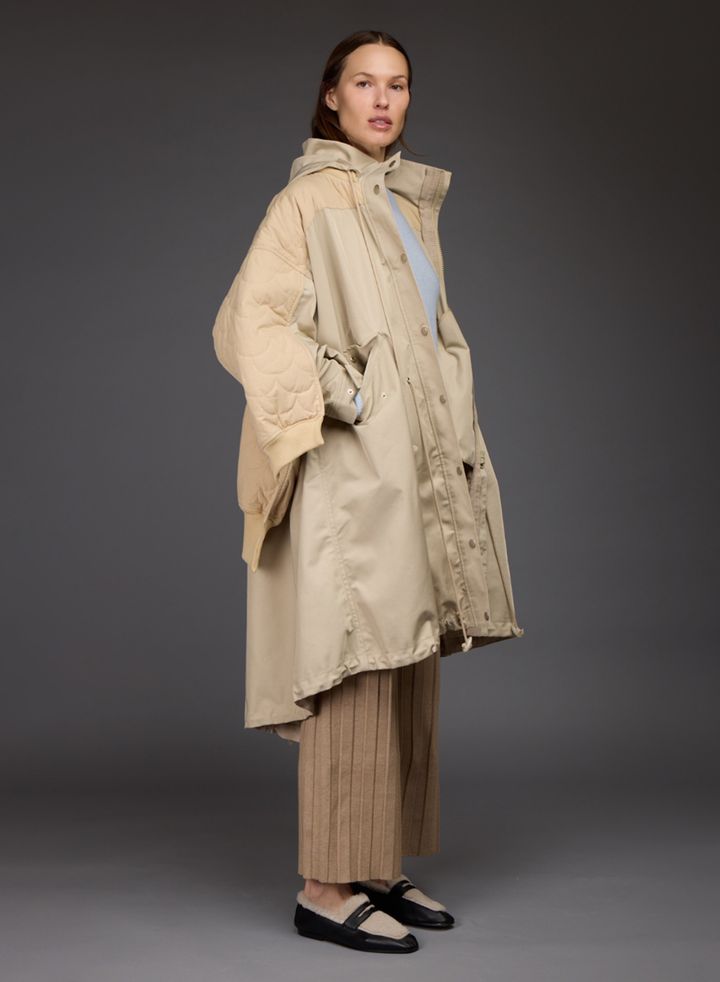 A model shown from the back wearing a beige quilted puffer jacket with a hood, a matching beige long coat, pleated beige trousers, and black and cream flats, set against a dark gray studio background.