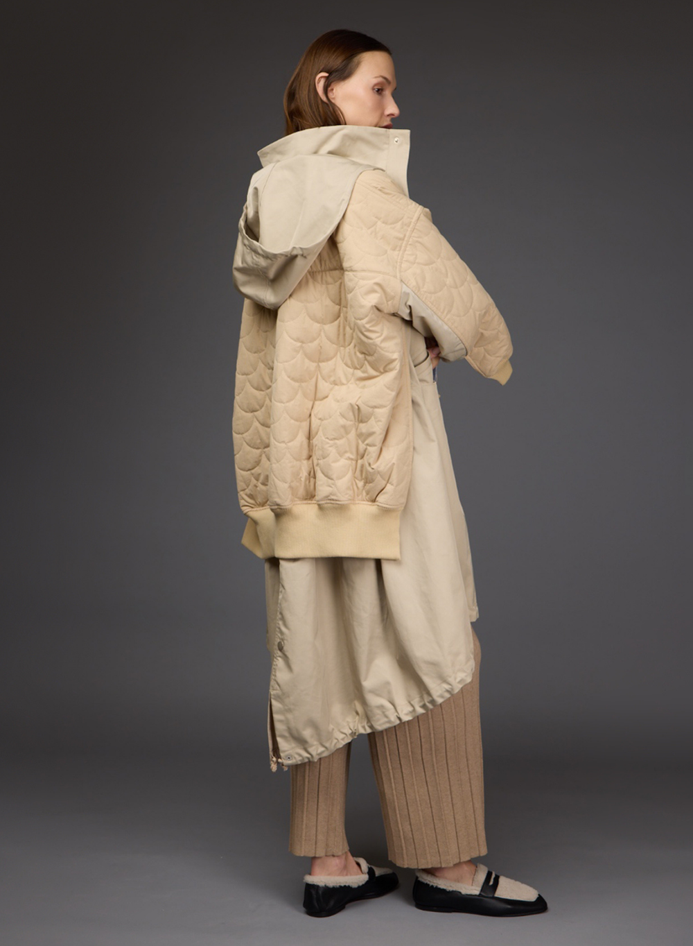 A model shown from the back wearing a beige Love Binetti quilted puffer jacket with a hood, a matching beige long coat, pleated beige trousers, and black and cream flats, set against a dark gray studio background.