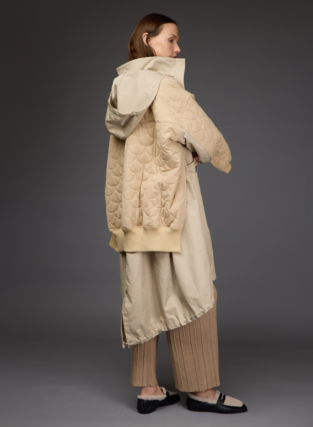 A model shown from the back wearing a beige Love Binetti quilted puffer jacket with a hood, a matching beige long coat, pleated beige trousers, and black and cream flats, set against a dark gray studio background.