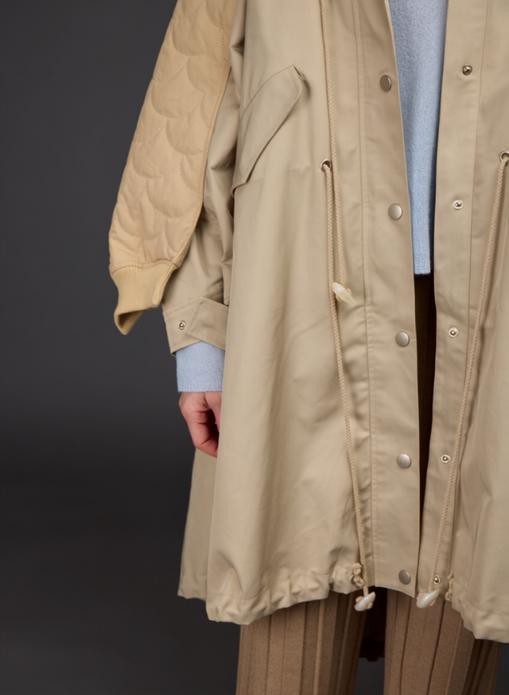 A close-up of a person wearing a beige hooded jacket with a quilted sleeve, a light blue knit sweater, and brown pleated trousers against a dark gray studio background.