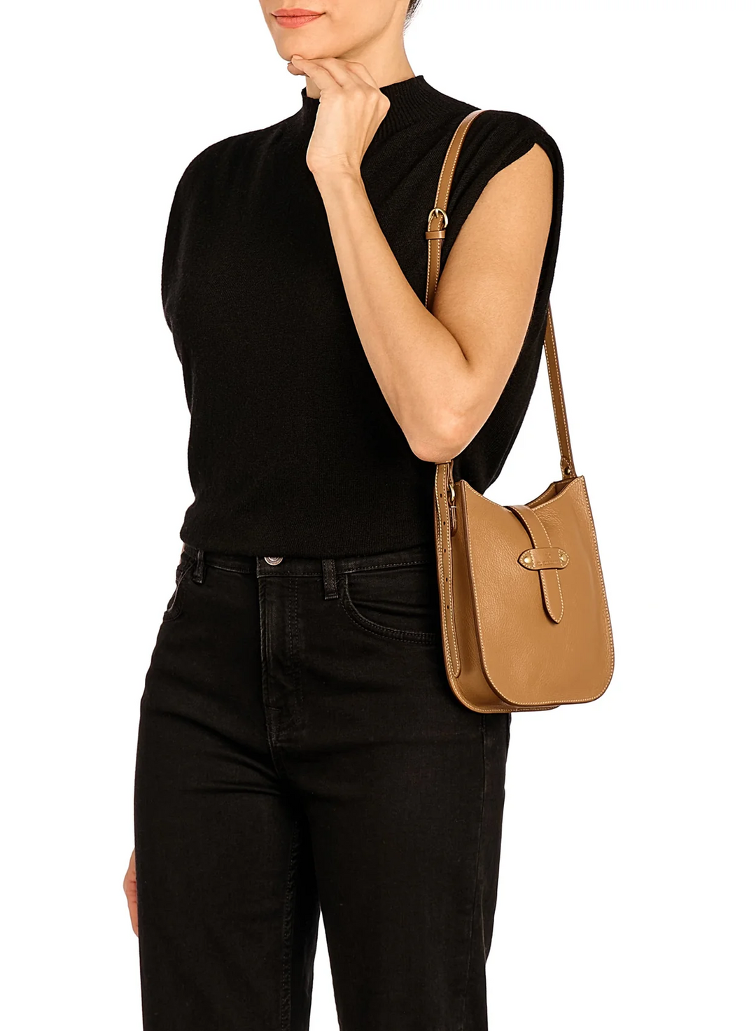 A woman wearing a black sleeveless mock-neck top and black jeans carries a tan leather shoulder bag with a front flap and buckle detail against a plain white background.