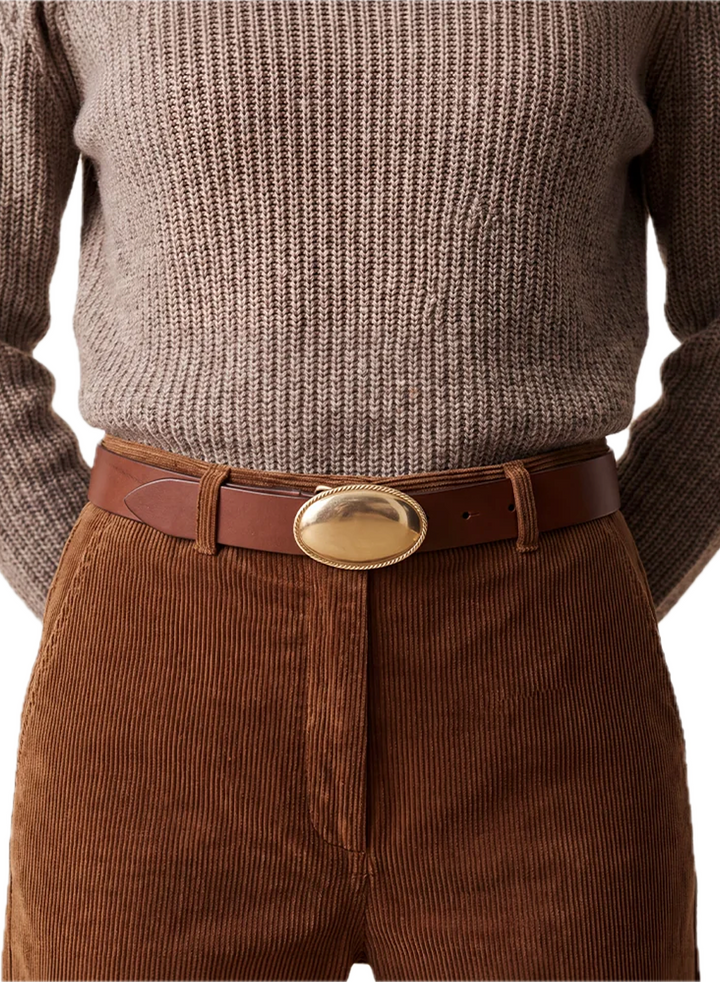 A close-up of a person wearing a brown leather belt with a large oval gold-tone buckle, styled with brown corduroy trousers and a taupe knit sweater.