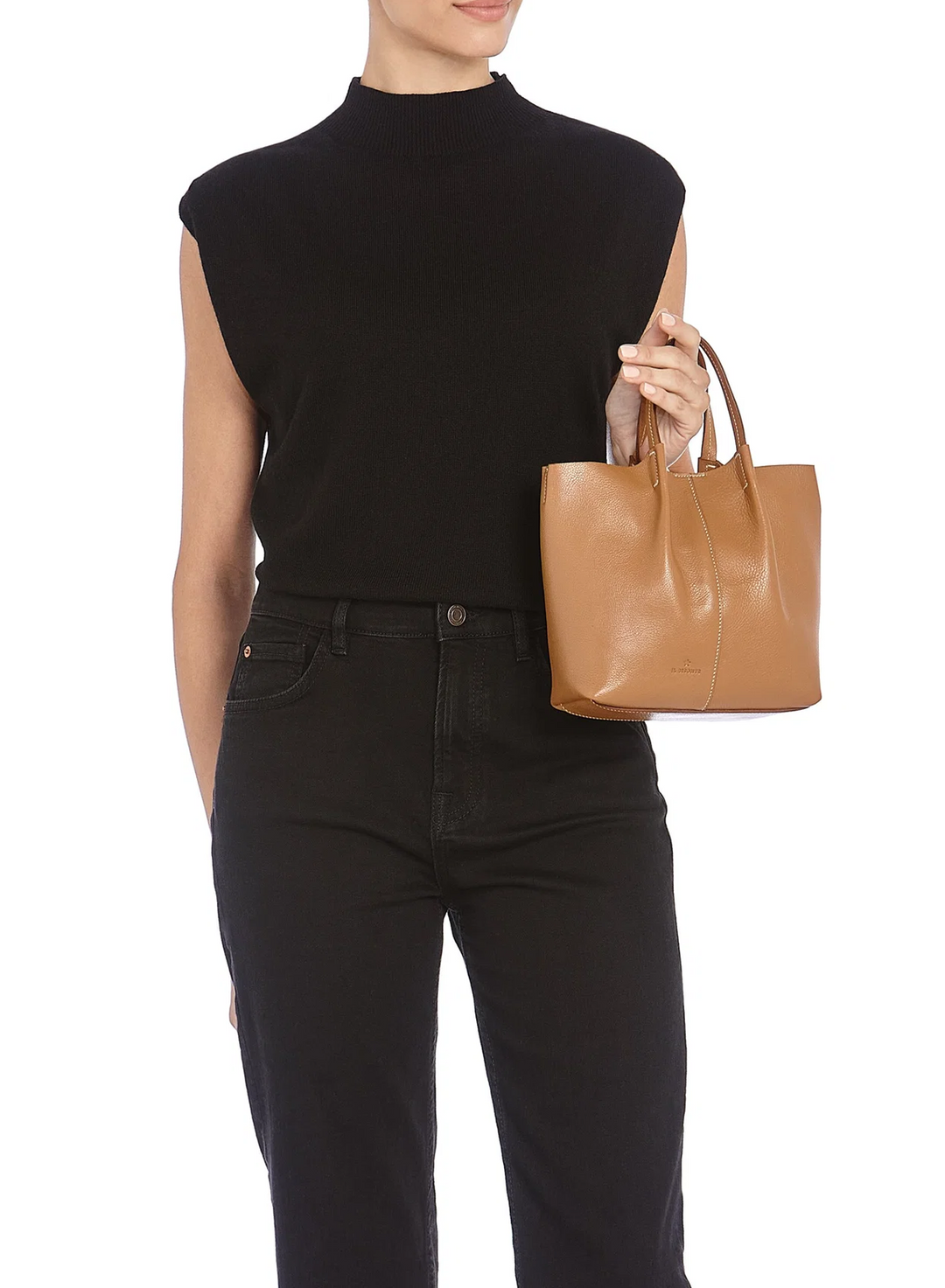 A model wearing a black sleeveless mock-neck top and black jeans holds a tan leather tote bag against a plain white background.
