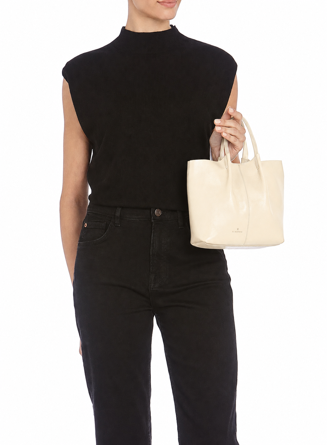 A model wearing a black sleeveless mock-neck top and black jeans, holding a cream-colored leather tote bag against a plain white background.