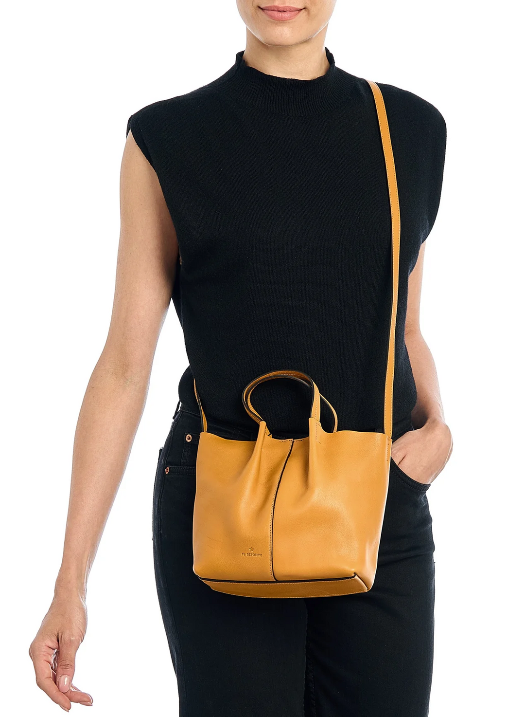 A woman wearing a black sleeveless mock-neck top and black pants, carrying a mustard yellow leather crossbody bag with top handles and a thin shoulder strap against a white background.