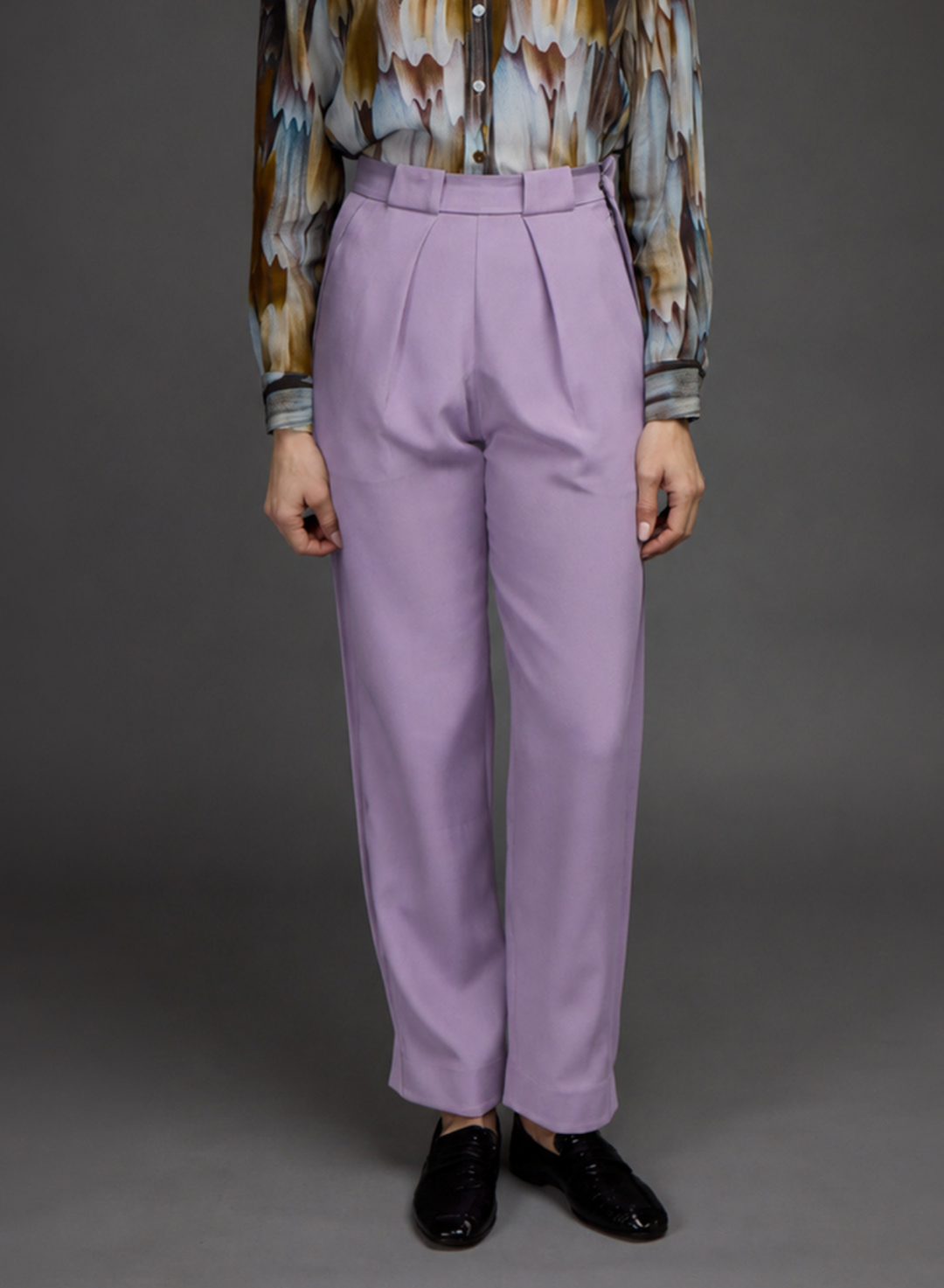 A model is shown from the chest down wearing lavender wide-leg pleated trousers, a patterned button-up shirt, and black loafers against a solid gray studio background.