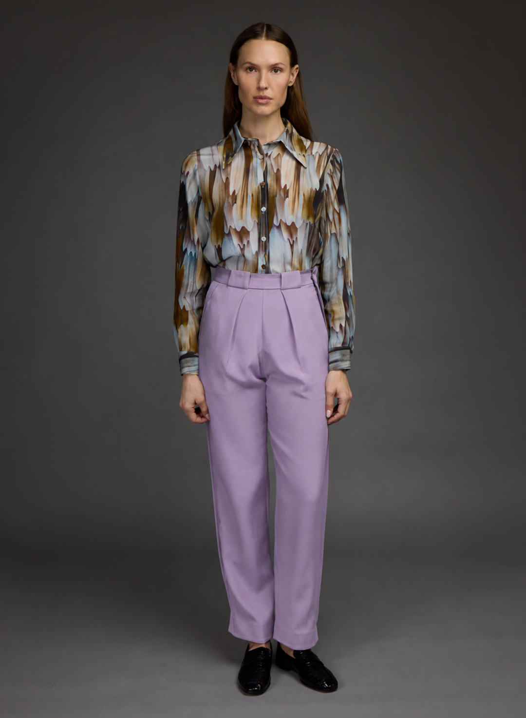 A model stands against a dark gray studio background wearing a long-sleeve button-up shirt with an abstract brown, blue, and beige pattern, paired with high-waisted lavender pleated trousers and black loafers.