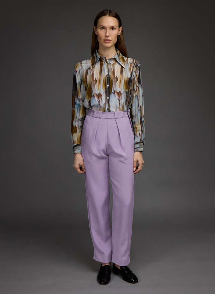 A model stands against a dark gray studio background wearing a long-sleeve button-up shirt with an abstract brown, blue, and beige pattern, paired with high-waisted lavender pleated trousers and black loafers.