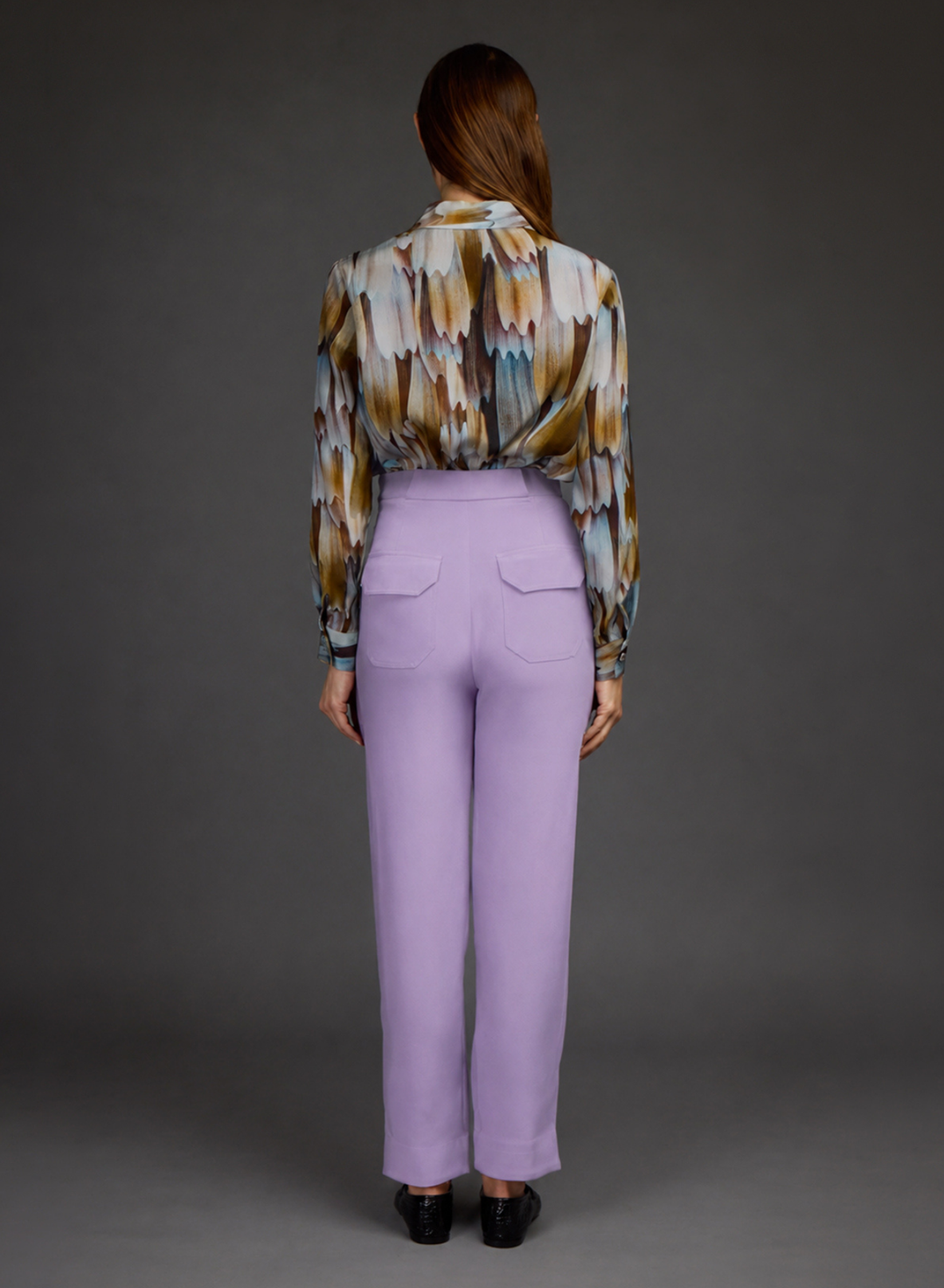A woman is shown from the back wearing a patterned long-sleeve blouse and lavender high-waisted trousers against a dark gray studio background.