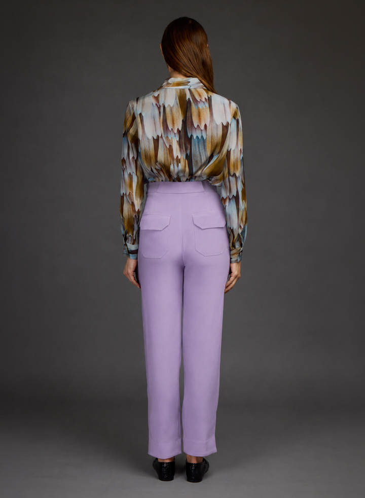 A woman is shown from the back wearing a patterned long-sleeve blouse and lavender high-waisted trousers against a dark gray studio background.