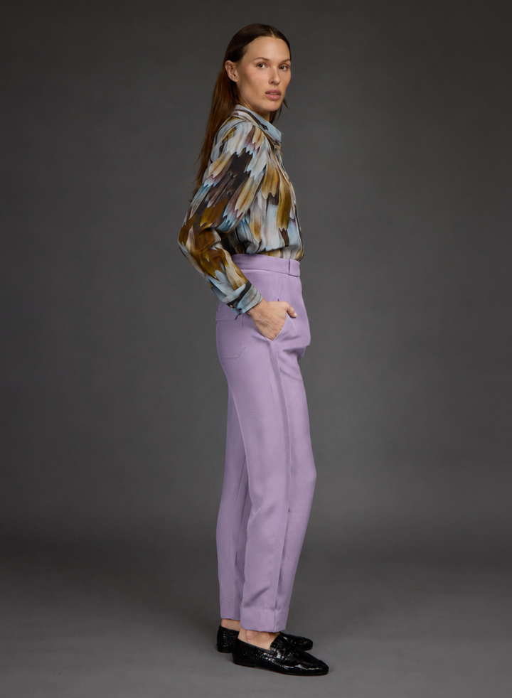 A woman stands in profile wearing a long-sleeve abstract print blouse in brown, beige, and blue tones, paired with high-waisted lavender trousers and black loafers, against a dark gray studio background.