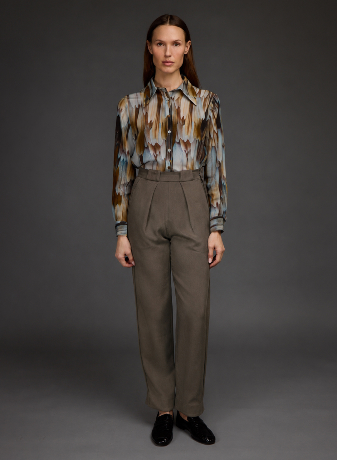 A model stands against a dark gray studio background wearing a long-sleeve button-up shirt with an abstract brown, blue, and beige pattern, paired with high-waisted taupe trousers and black loafers.