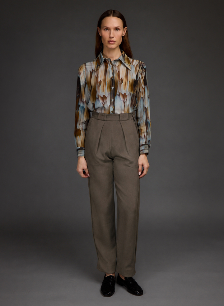 A model stands against a dark gray studio background wearing a long-sleeve button-up shirt with an abstract brown, blue, and beige pattern, paired with high-waisted taupe trousers and black loafers.