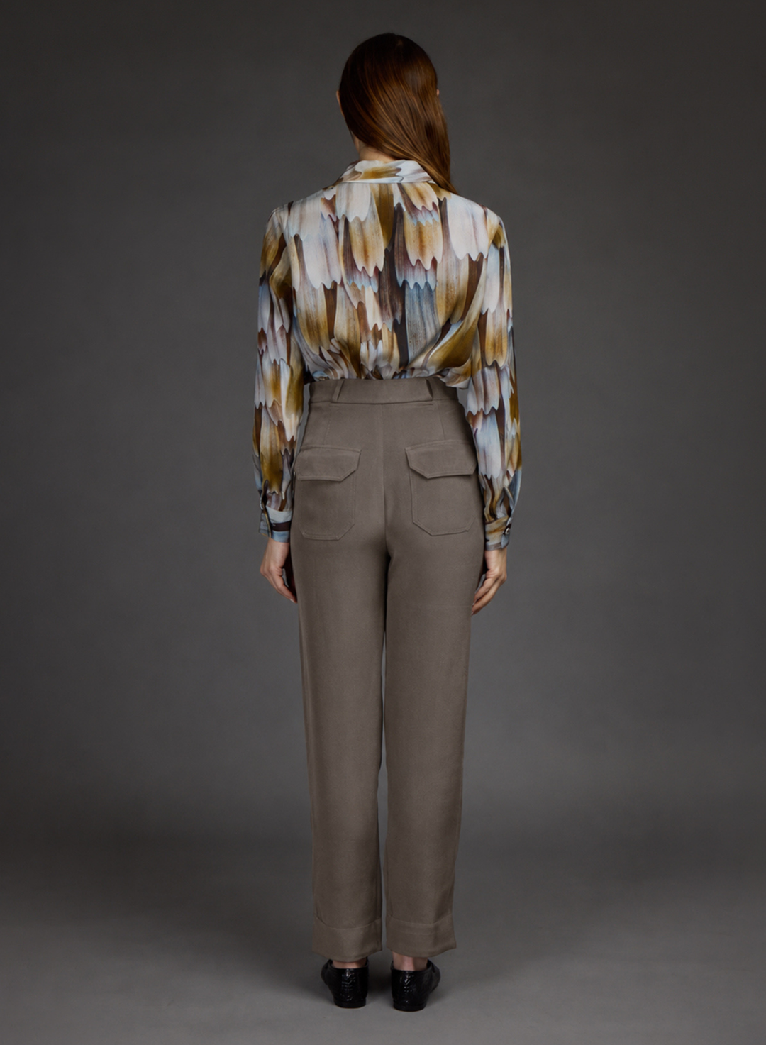 A model viewed from the back wearing a long-sleeve button-up shirt with a multicolor feather-like pattern in beige, brown, white, and blue, paired with high-waisted taupe trousers and black shoes, set against a dark gray studio background.