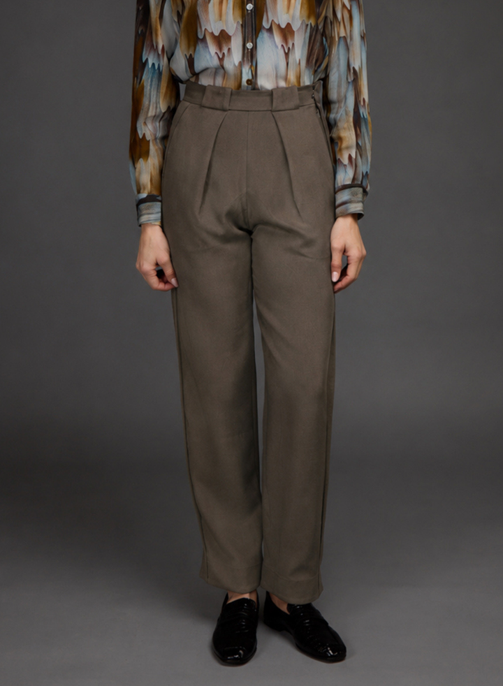 A model is shown from the chest down wearing high-waisted, wide-leg taupe trousers, a patterned long-sleeve shirt, and black leather loafers against a dark gray studio background.