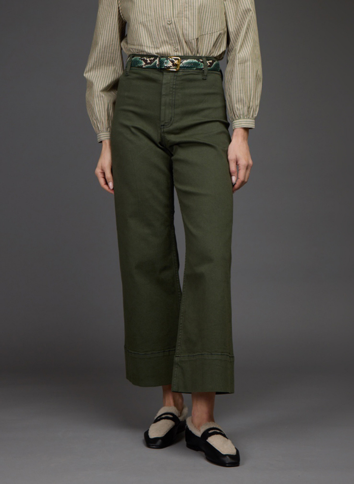 A model is shown from the waist down wearing olive green wide-leg cropped trousers, a beige and white striped shirt, a patterned belt, and black-and-beige loafers, set against a solid dark gray studio background.