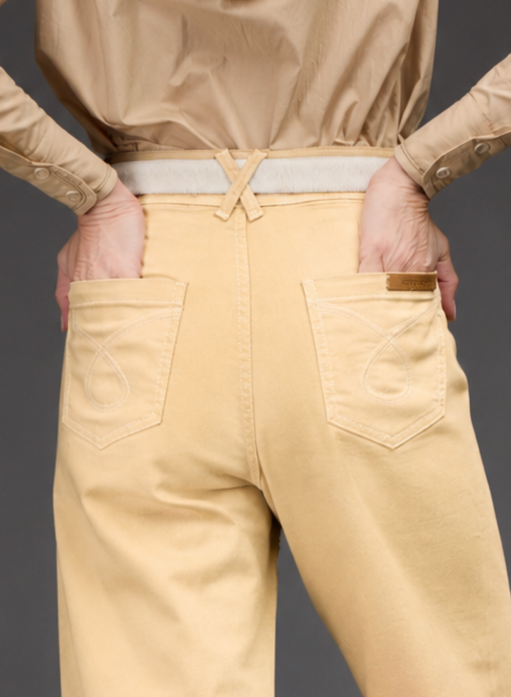 Close-up back view of a person wearing light beige trousers with back pockets, a matching belt, and a tan jacket against a dark gray background.
