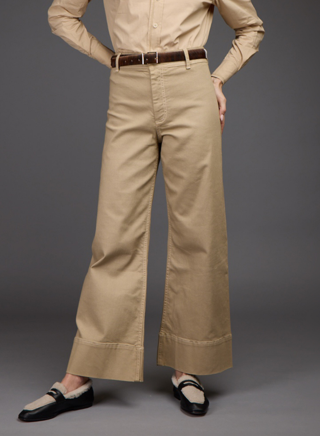 A model is shown from the waist down wearing beige wide-leg trousers with a brown belt, paired with a beige shirt and black-and-beige loafers, against a solid gray studio background. 