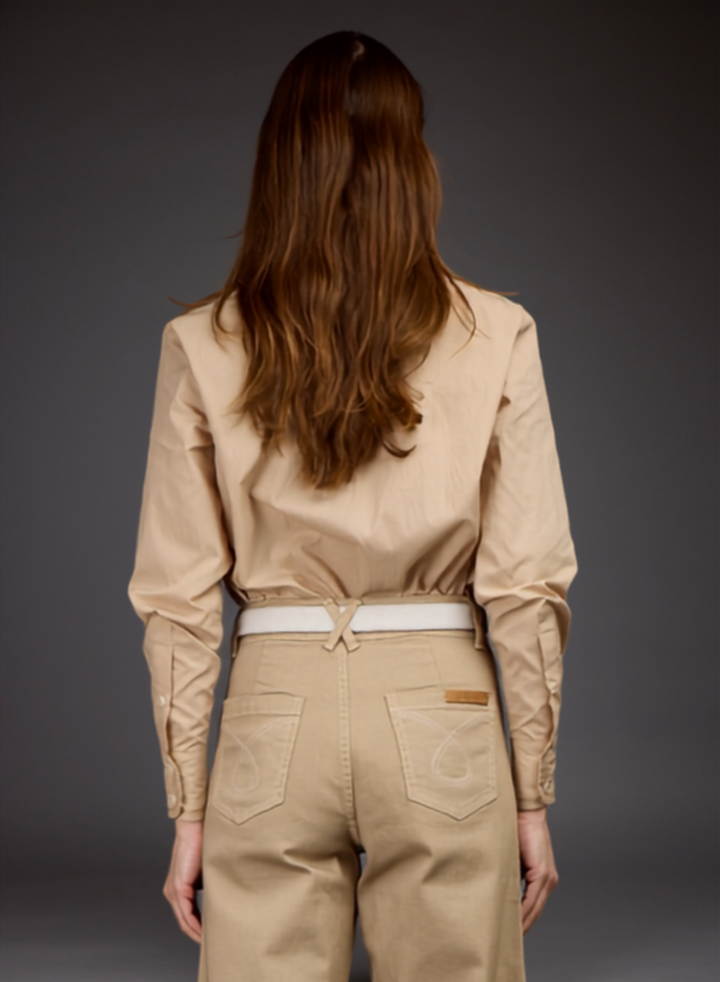 A woman viewed from behind wearing a beige long-sleeve button-up shirt tucked into matching beige pants with a white belt, against a dark gray studio background.