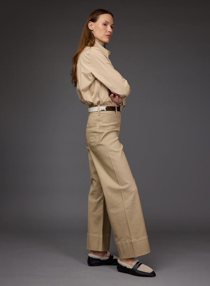 A woman stands in profile wearing a beige long-sleeve button-up shirt tucked into matching beige wide-leg trousers, with a thin brown belt and black slip-on shoes, against a dark gray studio background.
