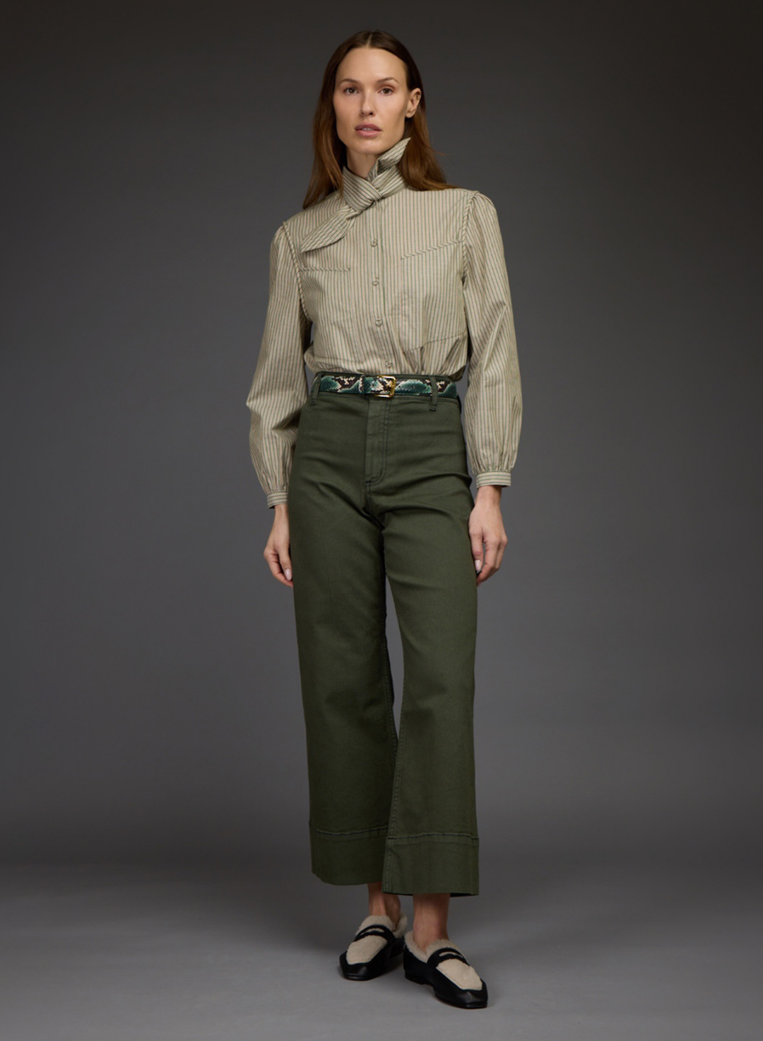 A model stands against a dark gray studio background wearing a beige and white vertically striped long-sleeve shirt with a high mock neck, paired with olive green cropped trousers and black Mary Jane shoes.