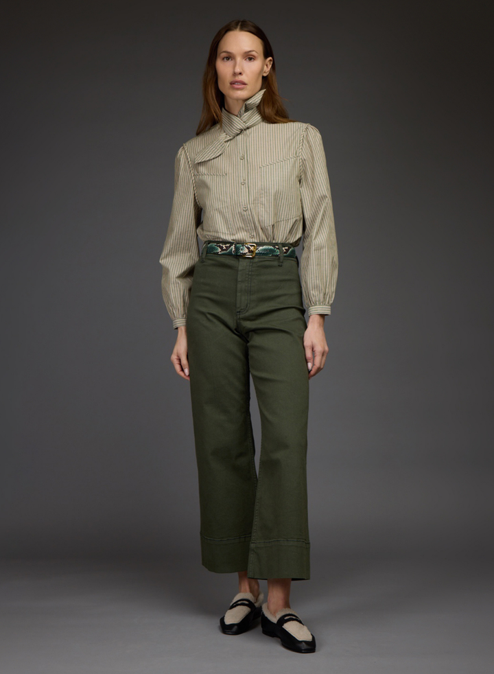 A model stands against a dark gray studio background wearing a beige and white vertically striped long-sleeve shirt with a high mock neck, paired with olive green cropped trousers and black Mary Jane shoes.