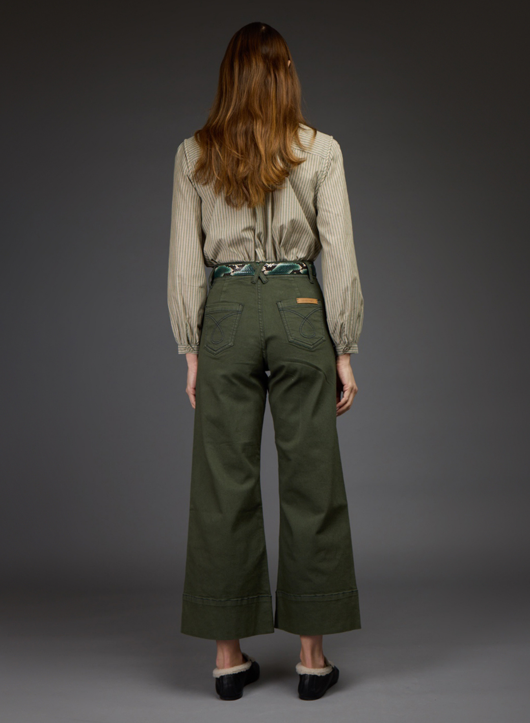 A woman viewed from the back wearing olive green wide-leg cropped pants, a beige ribbed long-sleeve blouse, and black shoes, standing against a dark gray studio background.